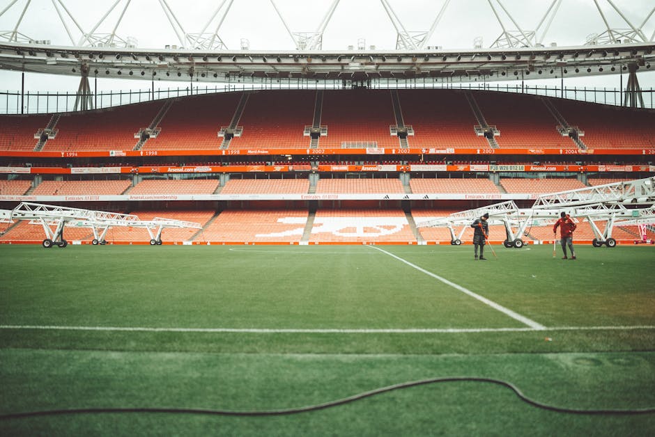The image shows the interior of Emirates Stadium in Islington, featuring a well-maintained football pitch with lush green grass and white boundary lines. The stadium's seating area, visible in the background, consists of red and orange seats arranged in tiers, and the upper tier has a curved roof structure supported by white steel beams. Two workers are present on the field, operating large scaffolding equipment used for maintenance or cleaning, with one worker dressed in red and the other in dark clothing. The lighting appears to be natural, indicating daytime, and the overall cleanliness of the field suggests professional maintenance. The scene exemplifies detailed surface cleaning and stadium maintenance, aligning with the professional cleaning services provided by Cleaner Islington, especially for large-scale venues like Emirates Stadium.