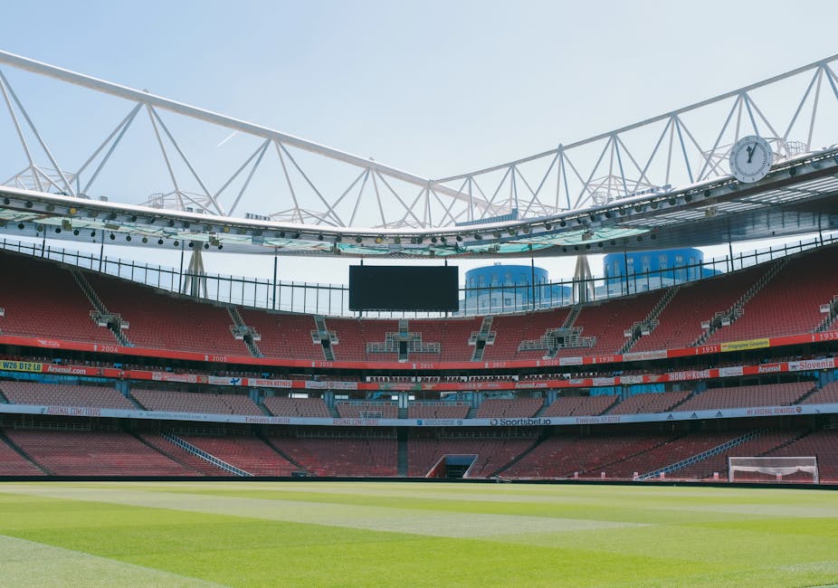 Empty stadium seating area with red and blue seats surrounding a well-maintained grass pitch. The structure includes a large overhead roof with white support trusses, and a digital scoreboard is visible in the background. Bright, natural daylight floods the stadium, highlighting the clean and orderly environment, typical of professional sports venues. This image reflects the importance of surface cleaning and maintenance to ensure hygiene and safety in such large-scale indoor and outdoor spaces. Cleaner Islington provides relevant deep cleaning and sanitisation services for stadiums and sports facilities to maintain their high standards of cleanliness and hygiene.