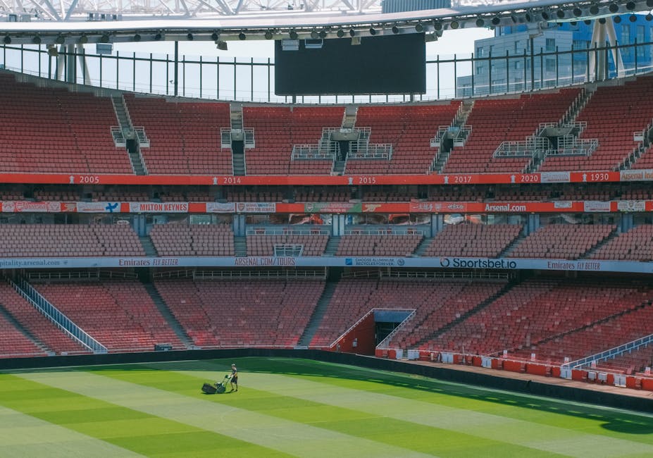 The image shows the interior of Emirates Stadium in Islington, featuring a well-maintained football pitch with lush green grass and white boundary lines. The stadium's seating area, visible in the background, consists of red and orange seats arranged in tiers, and the upper tier has a curved roof structure supported by white steel beams. Two workers are present on the field, operating large scaffolding equipment used for maintenance or cleaning, with one worker dressed in red and the other in dark clothing. The lighting appears to be natural, indicating daytime, and the overall cleanliness of the field suggests professional maintenance. The scene exemplifies detailed surface cleaning and stadium maintenance, aligning with the professional cleaning services provided by Cleaner Islington, especially for large-scale venues like Emirates Stadium.