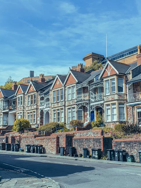 A row of Victorian-style terraced houses with decorative white trim and bay windows, situated on a slightly elevated street in Highbury Fields, Islington. The brick facades are clean and well-maintained, with some greenery and small bushes in front gardens. The pavement is clear, and trash bins are lined up along the curb. The sky above is bright blue with a few scattered clouds, indicating a sunny day. The scene suggests a quiet residential area, and the cleanliness and upkeep reflect regular domestic or commercial cleaning efforts, consistent with services provided by Cleaner Islington, as highlighted on their website page for deep flat cleaning.