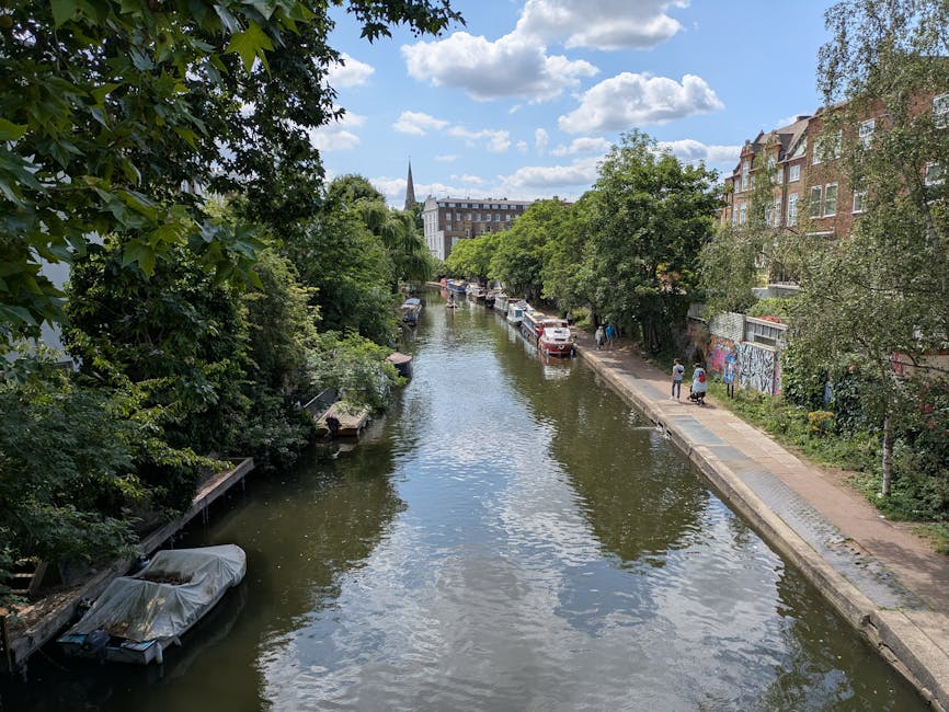 A tranquil canal scene in Highbury Fields, Islington, under a partly cloudy sky. The canal is bordered by a stone walkway on one side, with pedestrians walking along the path. Several boats are moored along the water's edge, with one covered by a protective tarp. Mature trees with lush green foliage overhang the canal, partially shading the walkway and creating a serene atmosphere. On the right, a row of residential buildings with brick facades and large windows stands adjacent to the promenade, some adorned with graffiti on a nearby wall. The water in the canal is calm, gently reflecting the sky and greenery, suggesting a peaceful urban environment. Overall, the scene epitomizes a well-maintained, scenic area characteristic of the Highbury Fields neighborhood, with an emphasis on natural beauty and community space, aligning with the professional cleaning and maintenance services provided by Cleaner Islington.