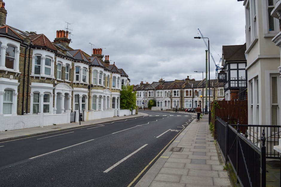 An empty residential street in Islington with Victorian-style terraced houses featuring white and brick facades, terracotta roof tiles, and decorative bay windows. The street has a paved sidewalk on the right with a black wrought-iron fence and a few small plants, and a tarmac road on the left with clear road markings. Overhead, the sky is cloudy, casting diffused natural light, highlighting the clean, well-maintained surfaces of the buildings and pavement. The scene reflects a quiet, tidy neighborhood with a focus on urban cleanliness, representative of the type of environment that Cleaner Islington services for end of tenancy and domestic cleaning, ensuring hygienic, polished finishes for both residential exteriors and interiors.