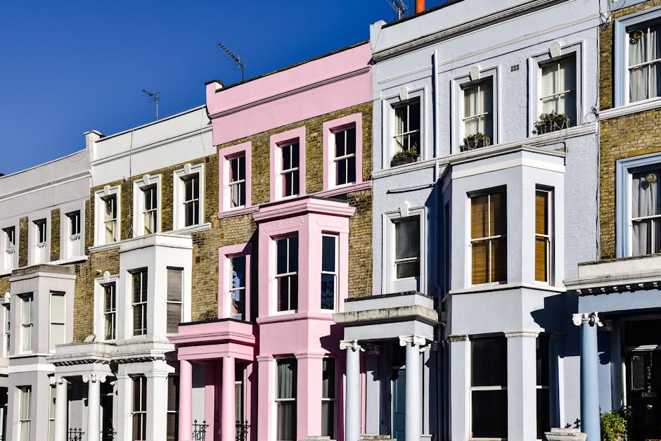 Photograph of a row of residential buildings with Victorian-style architecture on Upper Street in Islington, featuring large sash windows, decorative bay fronts, and a mix of brickwork and painted exteriors in white, pink, and pastel colors. The buildings are illuminated by natural daylight, highlighting their clean, well-maintained facades. Clear blue sky is visible above, and some windows have flower boxes or curtains visible inside. The overall appearance suggests a tidy, well-kept street scene, exemplifying pristine and hygienic urban residential environments, as maintained by professional cleaning services such as Cleaner Islington.