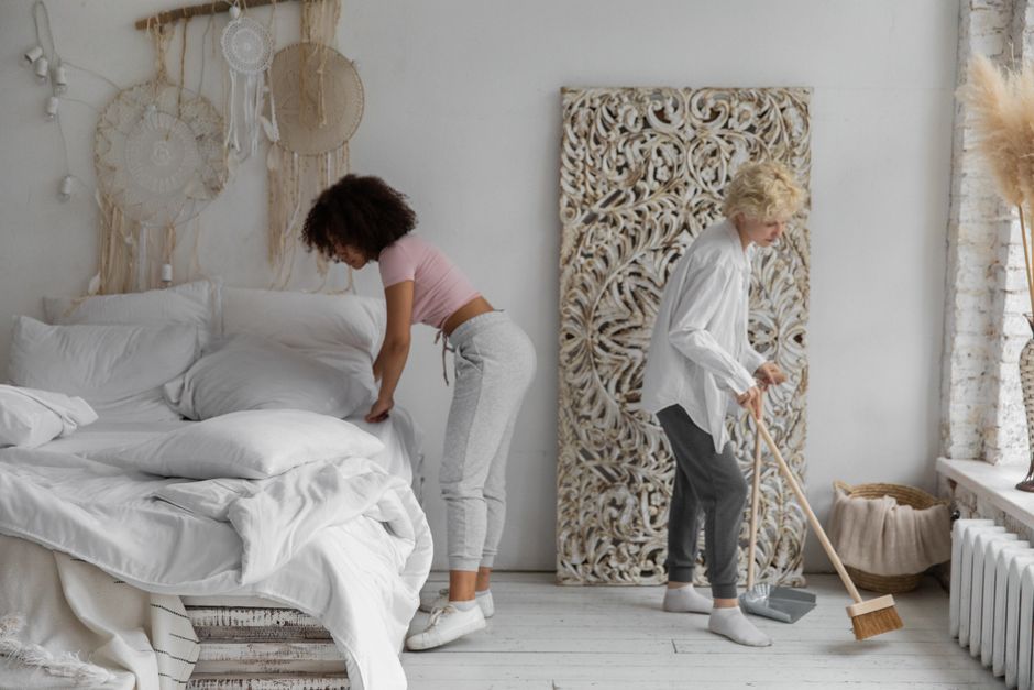 A woman with curly brown hair wearing a pink t-shirt and light gray sweatpants is tidying a white bed with rumpled linens in a bright bedroom. An older woman with short, curly blonde hair dressed in a white blouse and dark gray pants is sweeping the white wooden floor with a broom. The room features a white brick wall, decorative hanging wall art made of woven natural materials, and intricate carved wooden panels. Natural light filters through a window, illuminating the tidy space with a soft, airy atmosphere, demonstrating surface cleaning and general domestic cleaning in a modern, minimalist style, with an emphasis on cleanliness and maintenance, as highlighted by Cleaner Islington.