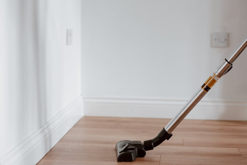 A handheld vacuum cleaner with a silver telescopic tube and black attachments resting on a light wooden floor near a white wall with baseboards. The vacuum is positioned diagonally, with the cleaning 