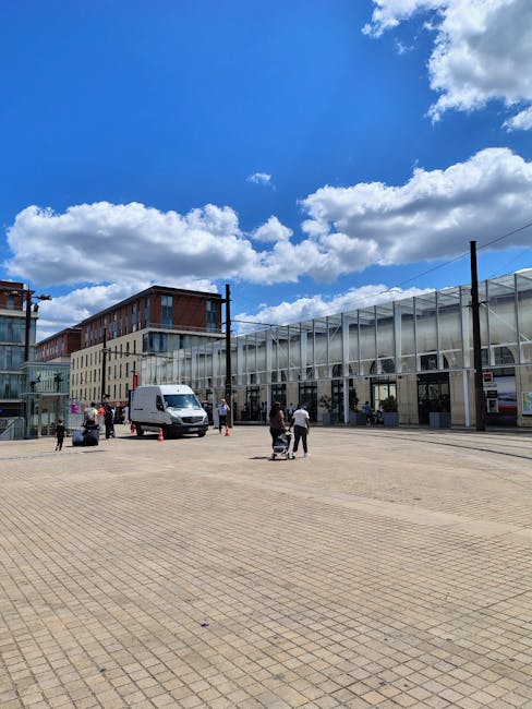 An outdoor urban scene featuring a wide paved plaza with a modern glass building in the background, under a bright blue sky with scattered white clouds. Several people are walking across the open space, and a white van is parked near the building. The building's façade has large glass panels and metallic structural supports, combining contemporary architectural materials like glass and steel. The scene is well-lit with natural sunlight, highlighting the clean surface of the paved area and the reflective surfaces of the building. This setting exemplifies a tidy, well-maintained public space typical of a clean, modern environment, as promoted by Cleaner Islington, which specializes in domestic and commercial cleaning services, including surface cleaning, deep cleaning, and sanitisation.