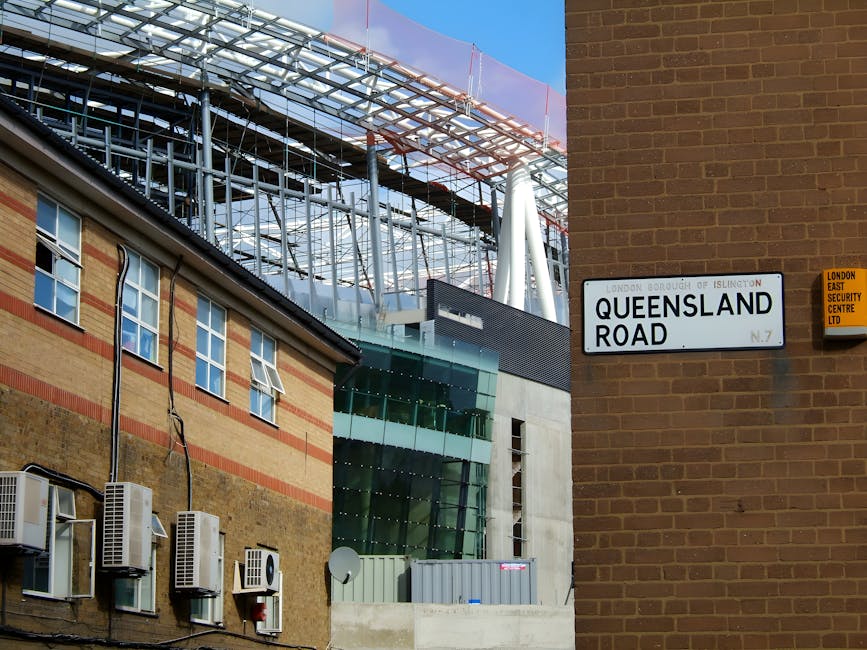A street scene on Queensland Road in Islington featuring a mix of architectural styles. In the foreground, there is a brick building with multiple air conditioning units mounted on the exterior wall. Adjacent to it is a modern glass and steel structure under construction, with visible scaffolding, reflective glass panels, and supporting beams. The street sign 'Queensland Road' is mounted on a brown brick wall on the right side of the image, alongside a yellow sign for London East Security Centre Ltd. Bright daylight illuminates the scene, highlighting the contrast between the older brick building and the new construction, with a clear blue sky in the background. This image reflects ongoing urban development and the need for professional domestic and commercial cleaning, as provided by Cleaner Islington, to maintain cleanliness amidst construction activity.